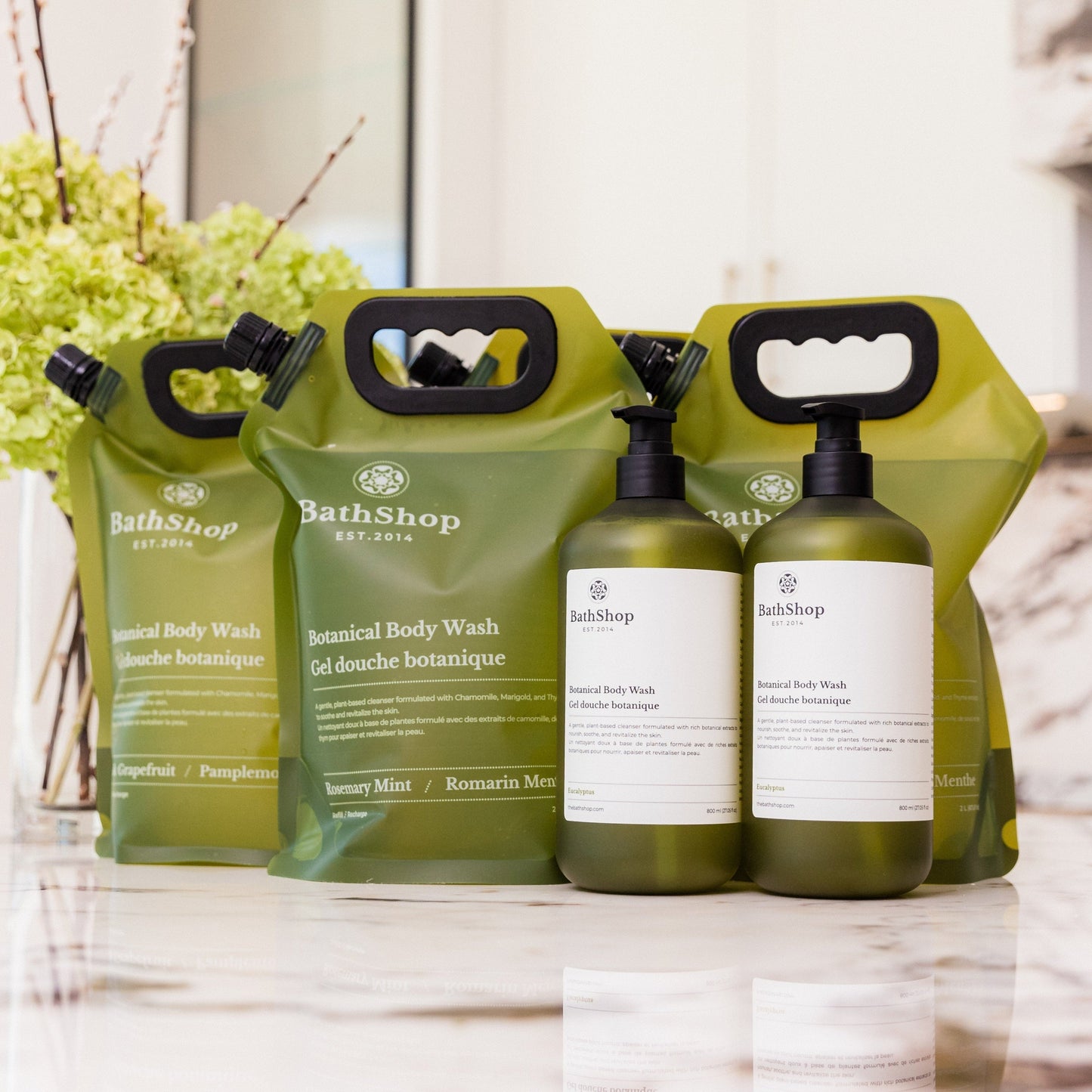 Plant-based and eco-friendly products by The Bath Shop including bottles and pouches on a marble surface with a blurred background.