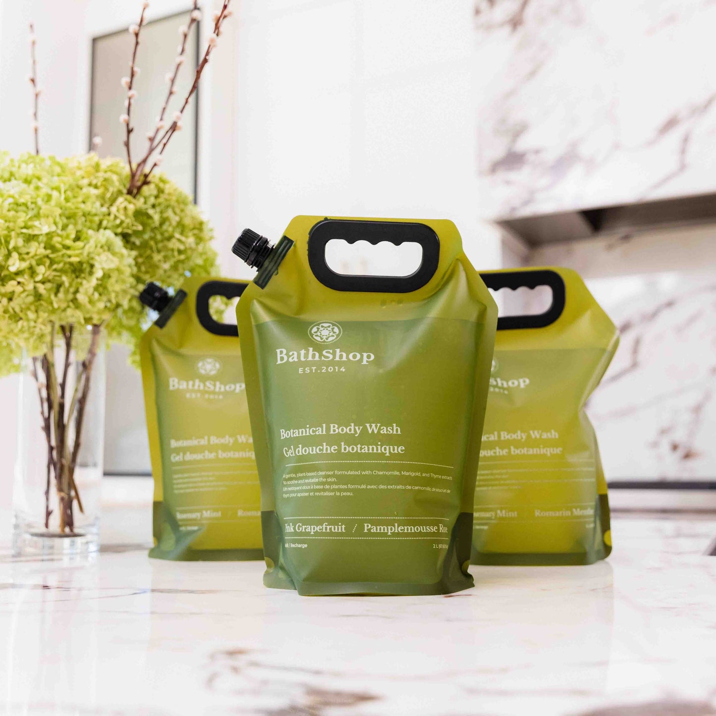 Three green refill pouches of BathShop Botanical Body Wash on a marble countertop with a decorative plant in the background.
