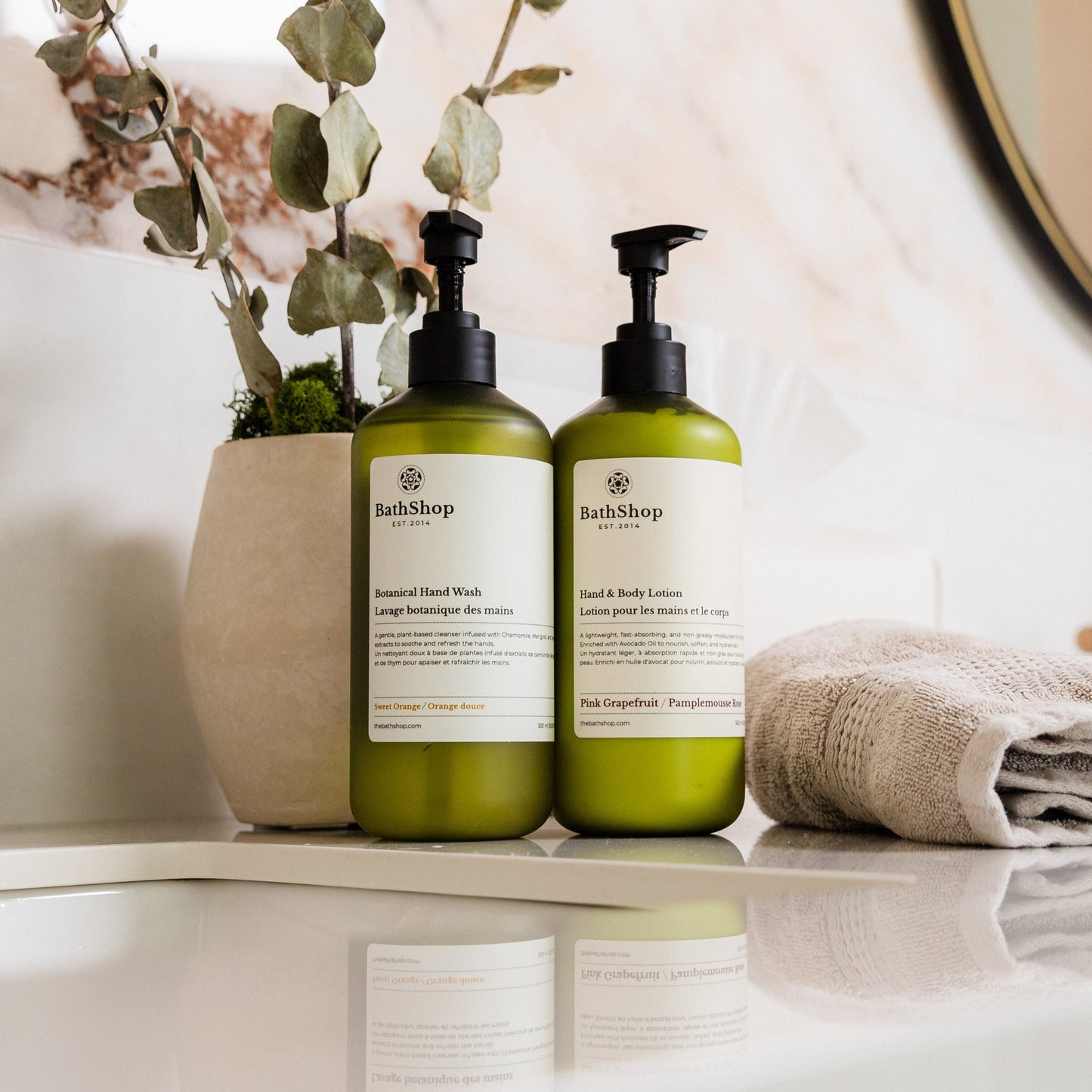 Two green bottles of The Bath Shop products on a bathroom shelf with a plant and towel in the background.